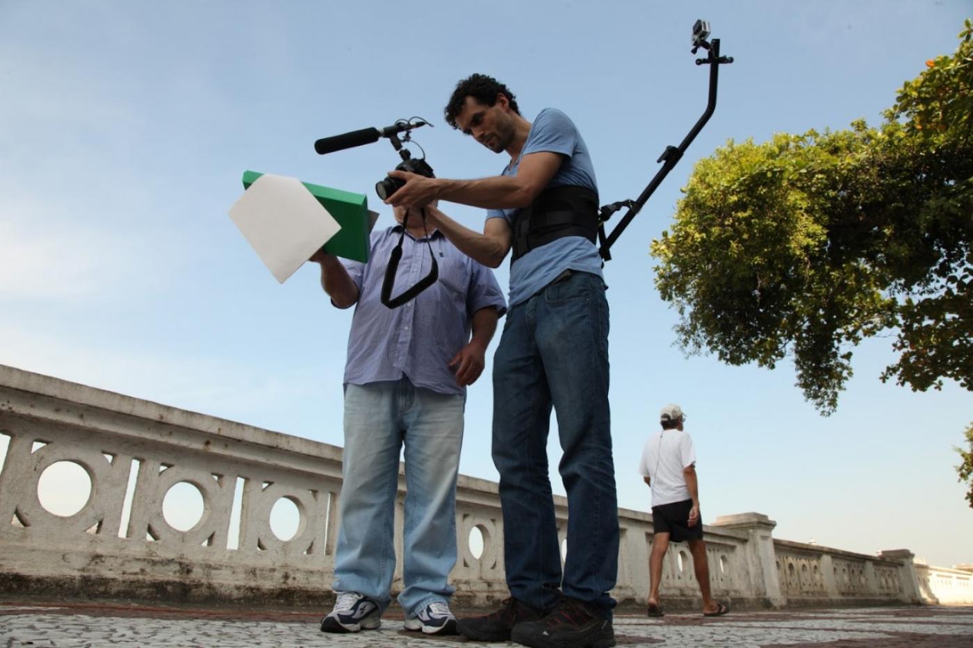 dois homens estão em pé no calçadão da orla diante de mureta. Eles estão fazendo uma gravação de vídeo. #paratodosverem