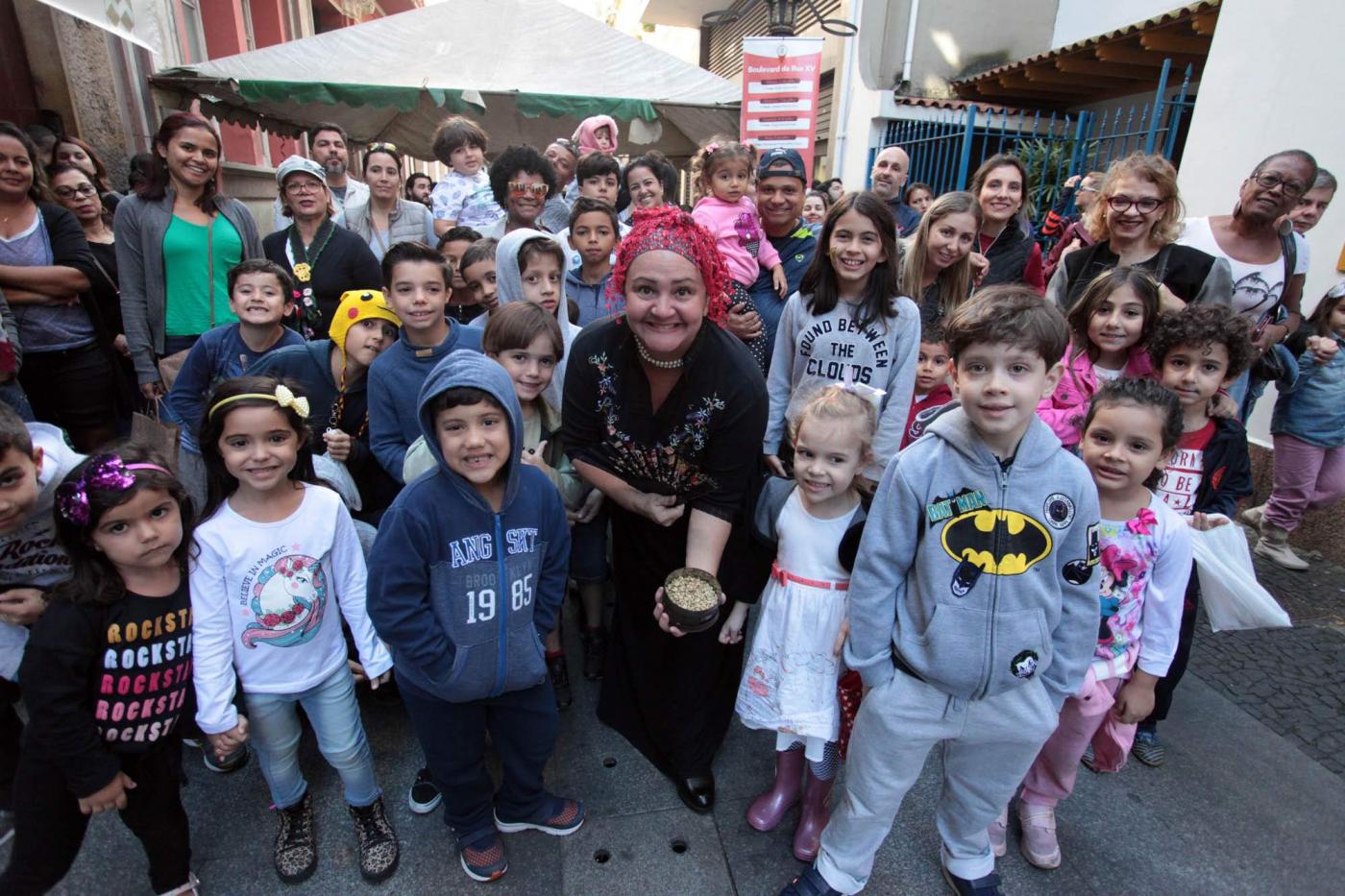 Crianças posam para foto junto a guia vestida com roupa típica portuguesa e uma lata com amostra de grãos de café. #Pracegover