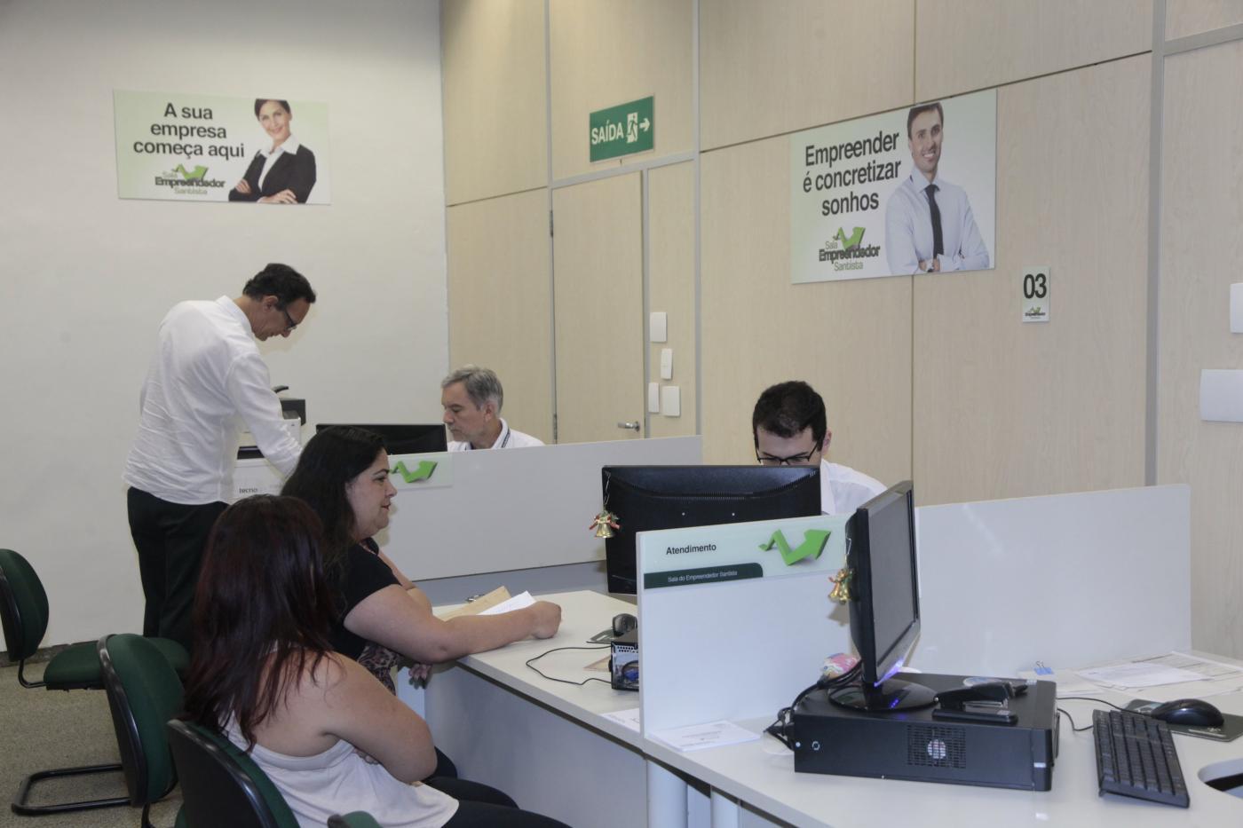 duas mulheres estão sendo atendidas por um homem. Elas de um lado da escrivaninha e ele do outro, em frente a um computador. #paratodosverem