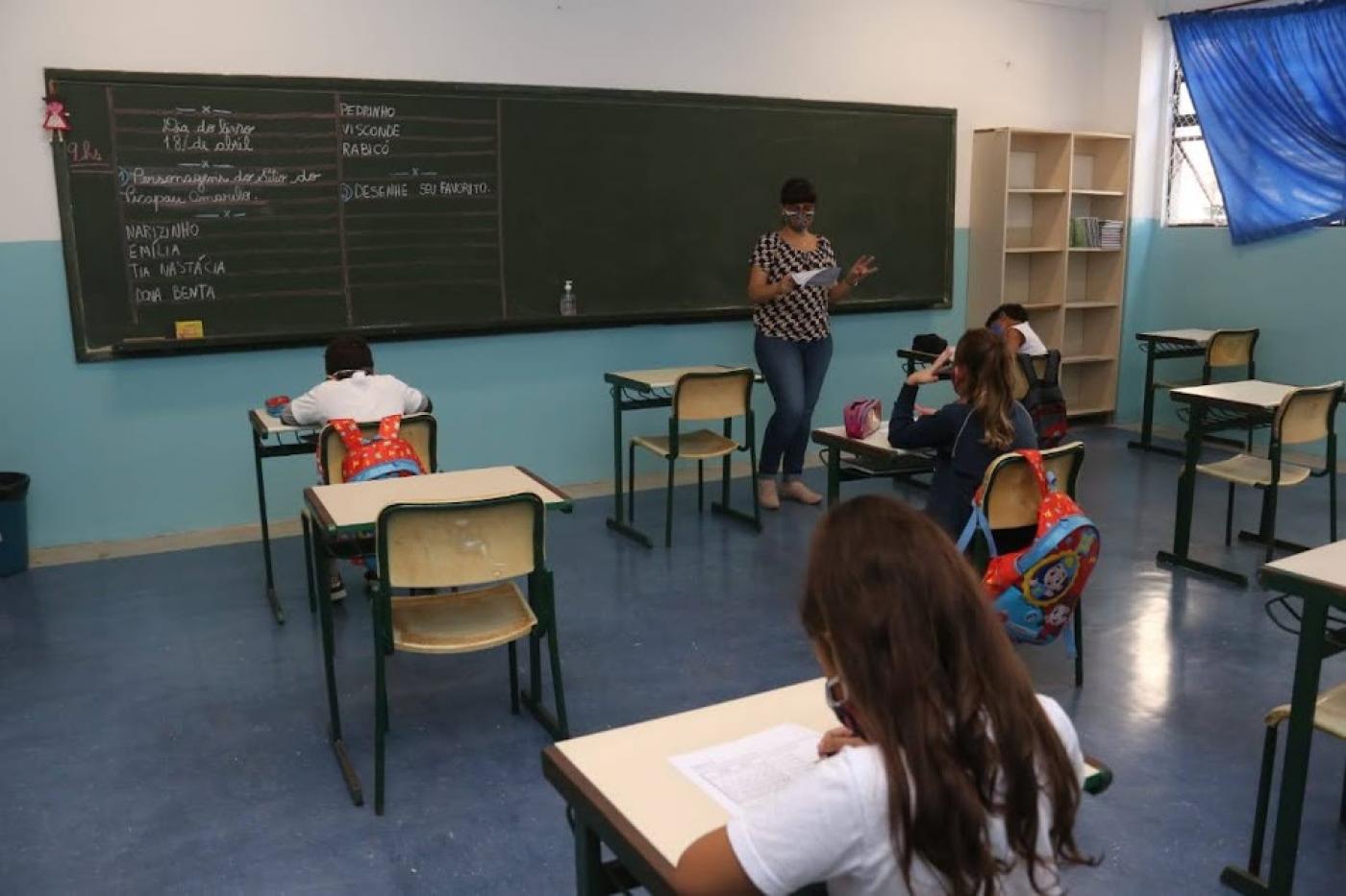 sala de aula com crianças sentadas, todas de costas. A professora está ao fundo, à frente da lousa e falando aos alunos. #paratodosverem