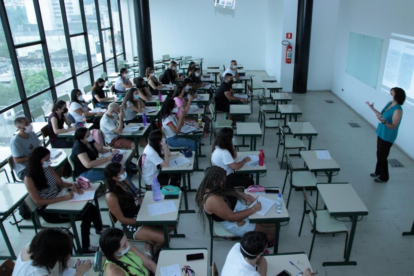 sala de aulas com alunos sentados e professora à frente. Sala é fechada por vidro que permite visualização de parte do centro histórico. #paratodosverem