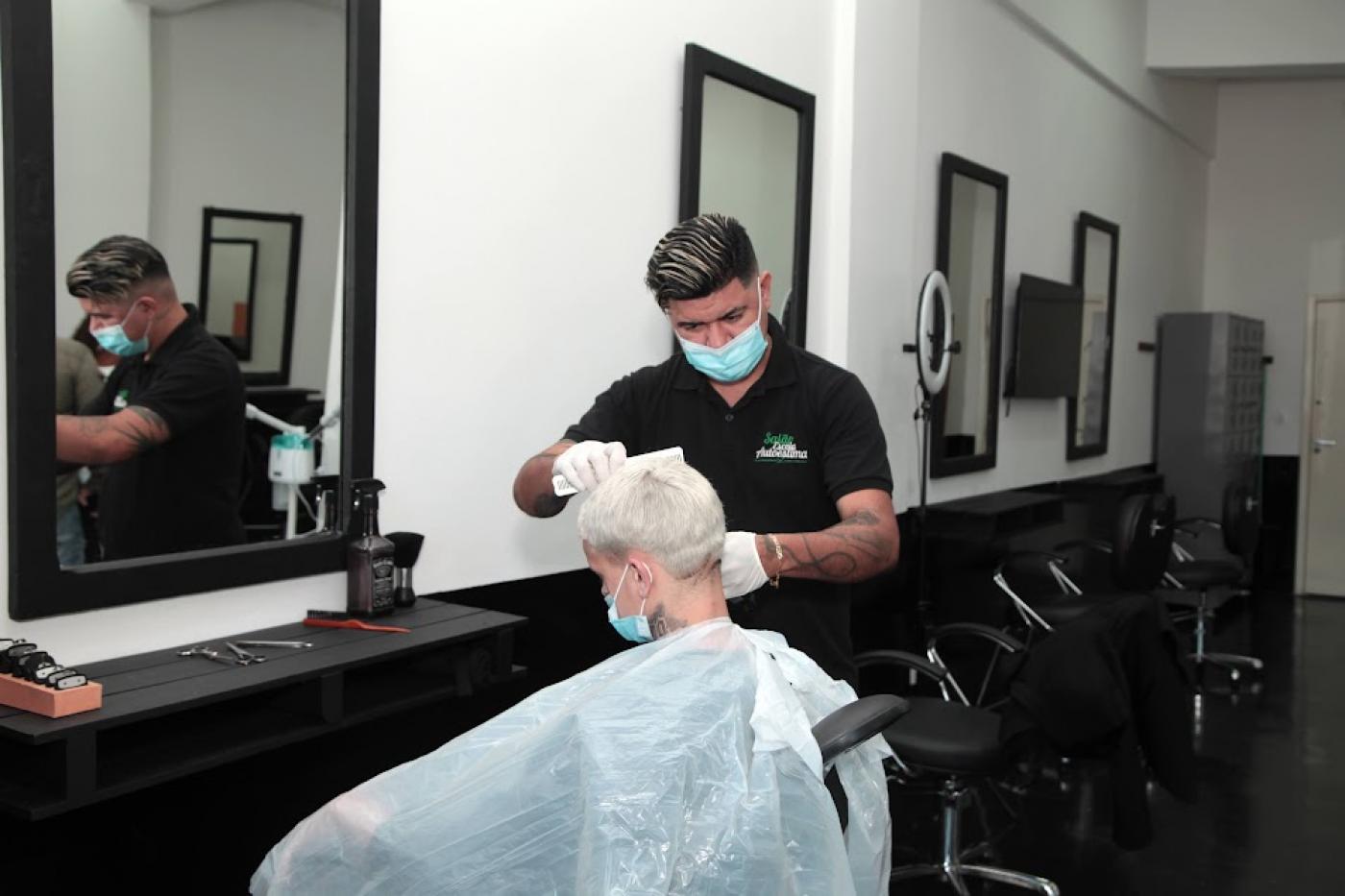 homem está em pé vestindo o uniforme preto do salão. Ele corta o cabelo de um rapaz que está sentado. O ambiente tem outras cadeiras para corte de cabelo. #paratodosverem