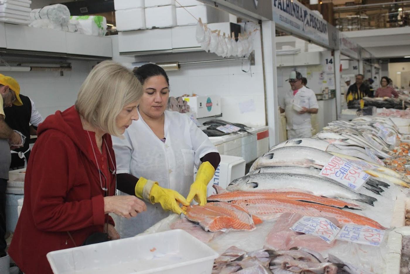 Cliente observa salmão exibido por profissional paramentada com luvas. #paratodosverem