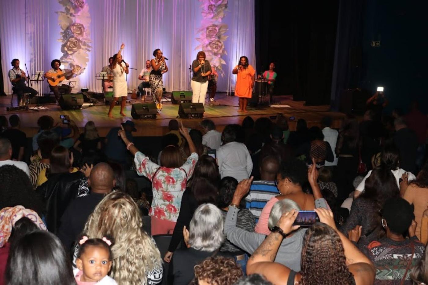 na plateia, público acompanha apresentação em pé, com mãos para o alto e alguns filmando com celular. #paratodosverem 