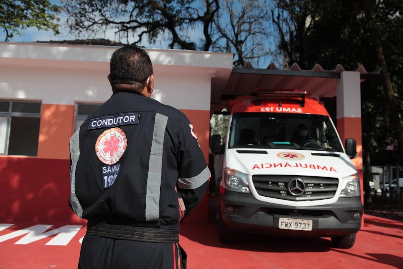 condutor uniformizado está de costas para a foto e de frente para a base do samu e para uma ambulância. #paratodosverem