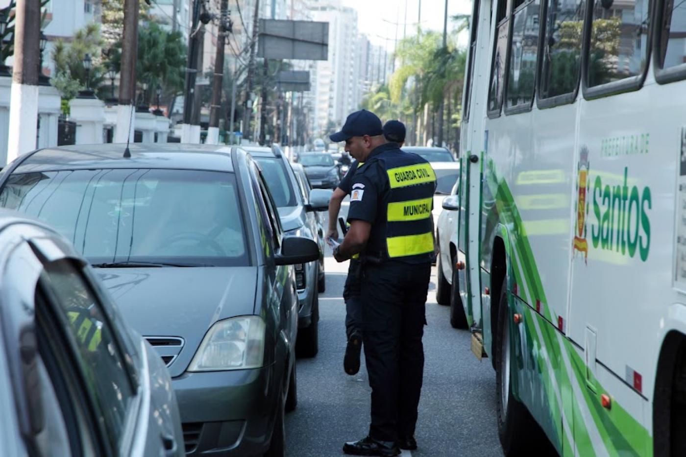 guardas municipais estão em pista de avenida, com carros parados. Eles estão entre carros e ônibus distribuindo folhetos. #paratodosverem