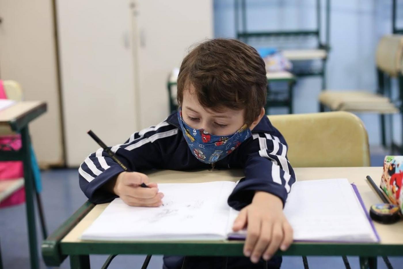 Criança de máscara escreve em um caderno em uma sala de aula #paratodosverem