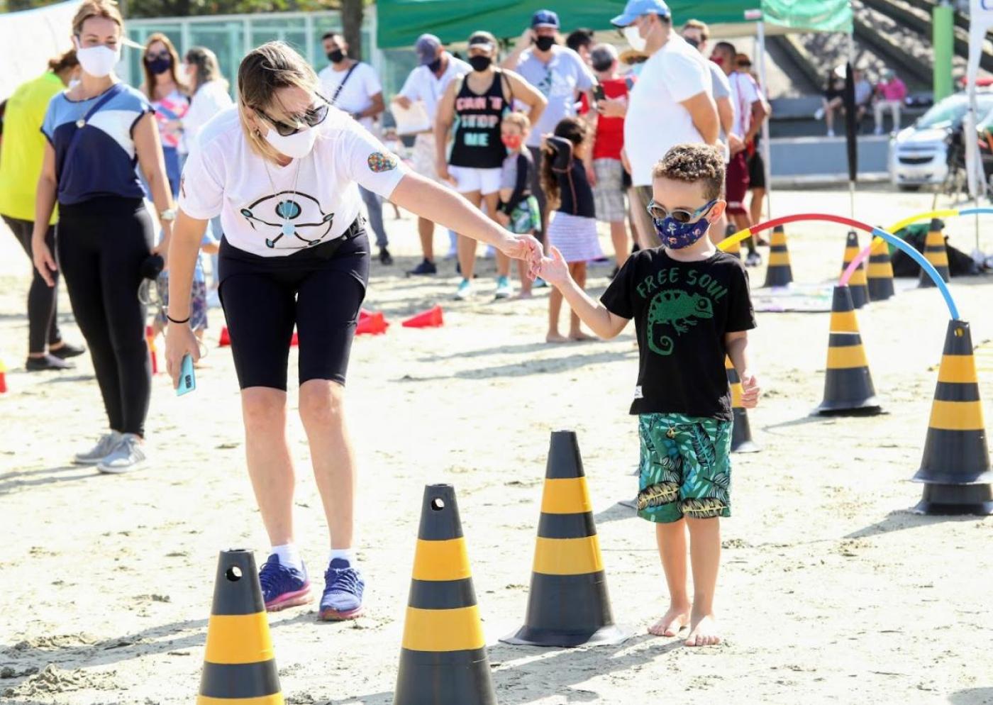 muilher segura mão de criança com deficiência visual, que é conduzida para caminhar entre cones na areia da praia. #paratodosverem