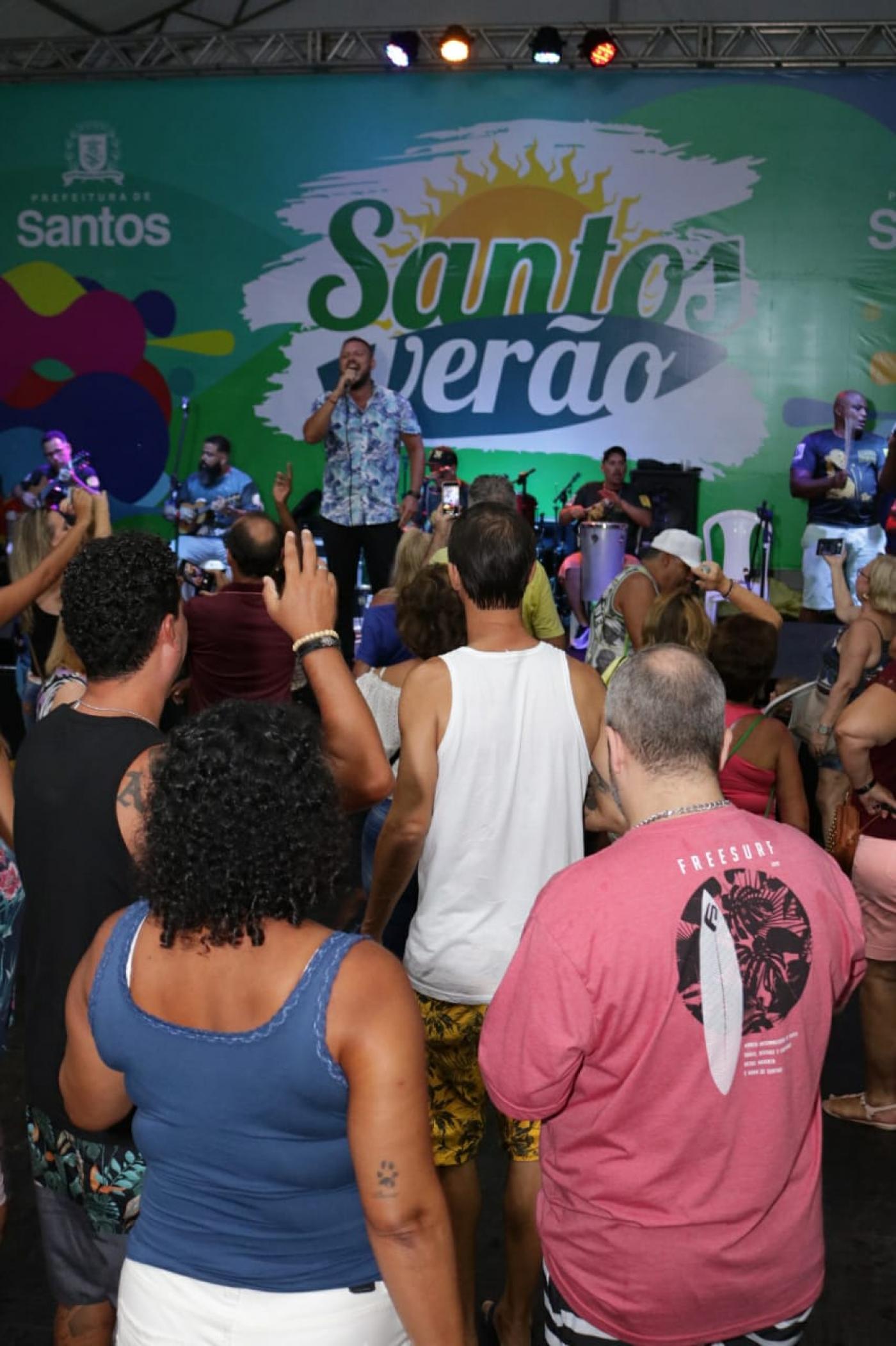 Público, de costas, assiste apresentação de cantor no palco com banner Santos Verão. #pracegover