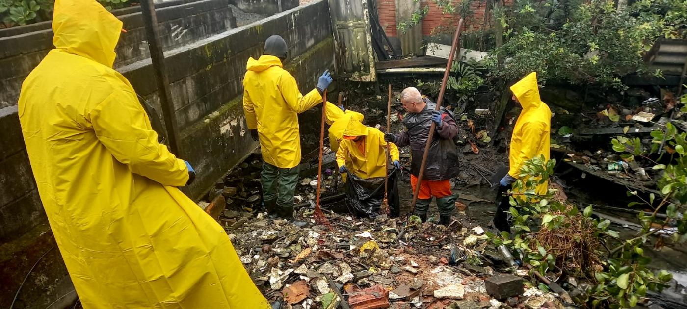 operários vestindo capas de chuva removem manualmente lixo de canal. #paratodosverem