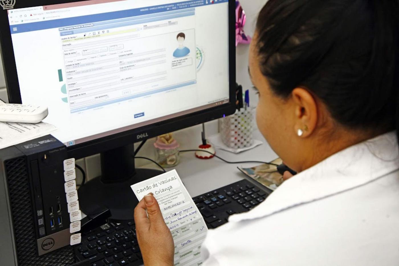 mulher segura carteira de vacinação diante de computador onde ela preenche dados. #paratodosverem