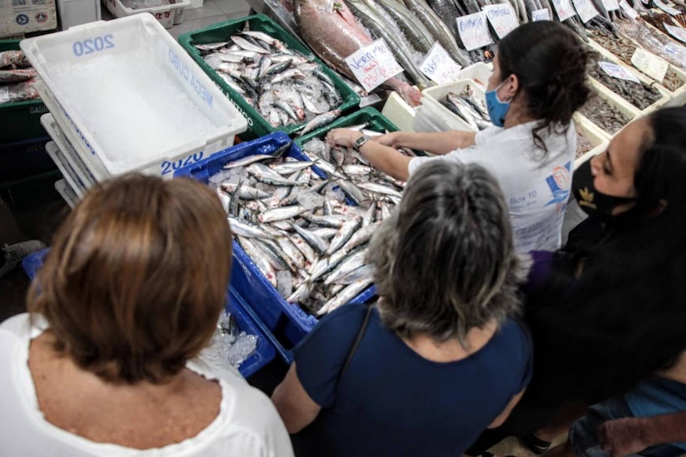 imagem do alto mostra mulheres à frente de balcão com caixas repletas de sardinhas. Uma delas, uniformizada, mexe em uma das caixas. #paratodosverem