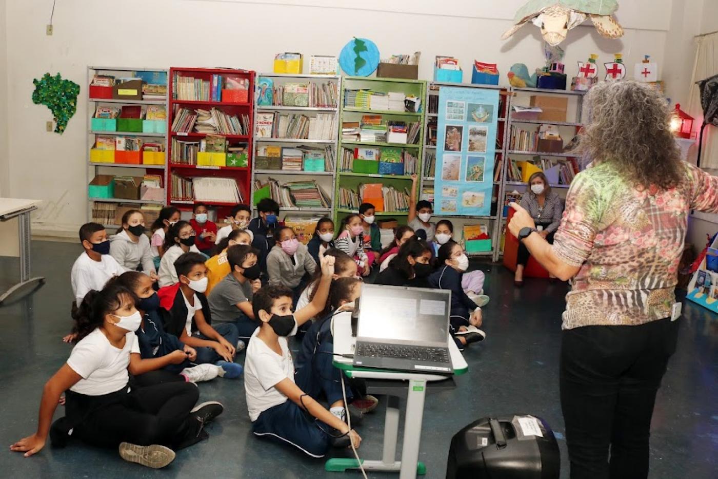 ALUNOS ESTÃO SENTADOS NO CHÃO ATENTOS A UMA MULHER QUE FALA EM PÉ. uM DELES LEVANTA A MÃO PARA PARTICIPAR. A sala tem prateleiras com livros e decoração voltada para o meio ambiente. #paratodosverem