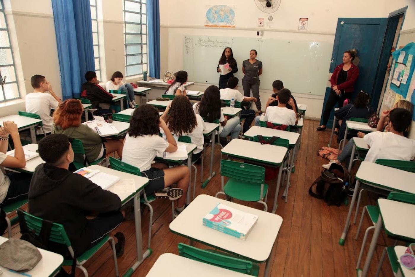 alunos estão tendo aula em sala. estão de costas para a foto. professores ao fundo em frente à lousa. #paratodosverem 