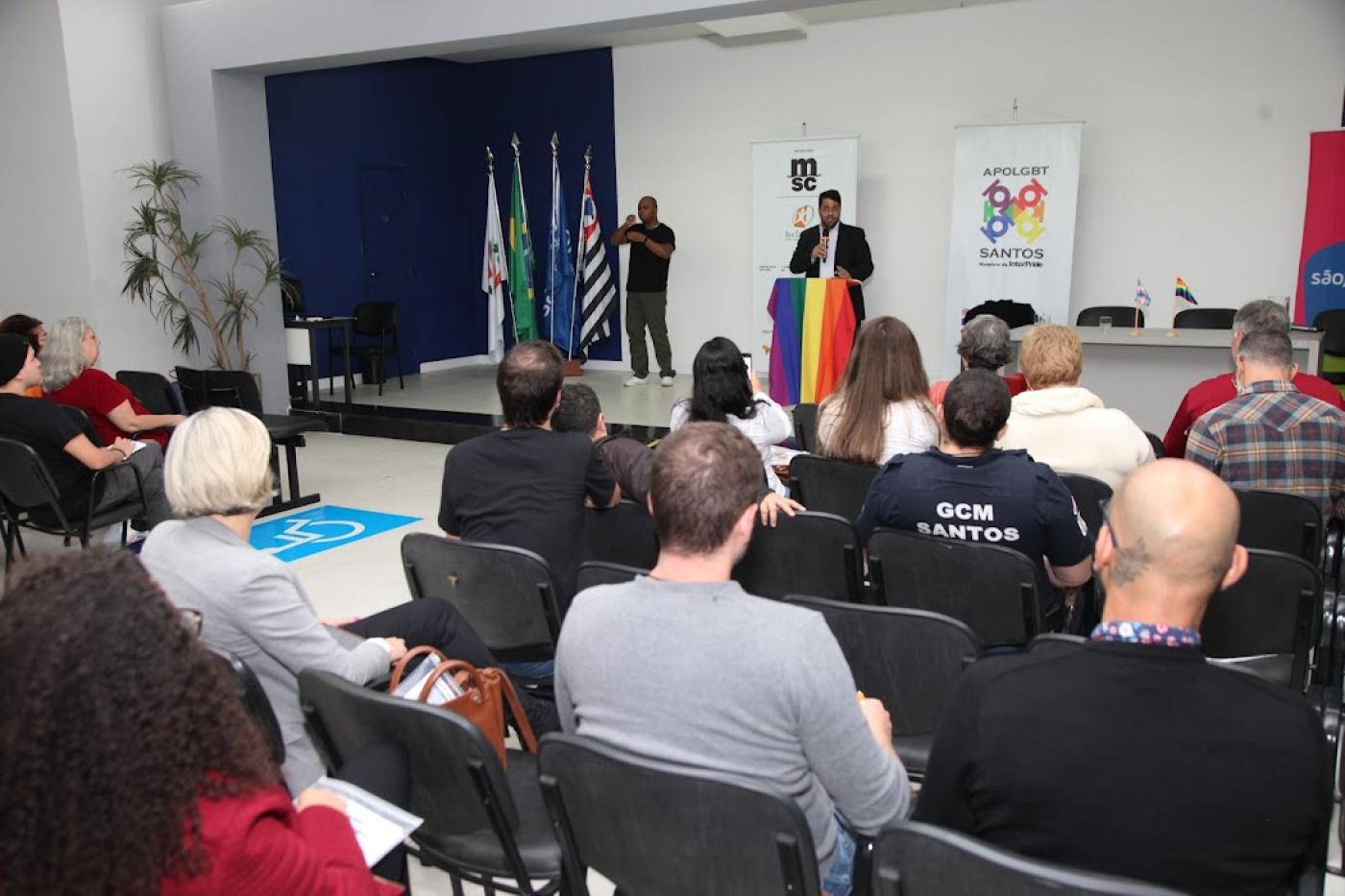 Palestrante em púlpito coberto com bandeira com as cores do arco-íris é observado pelo público sentado. #paratodosverem