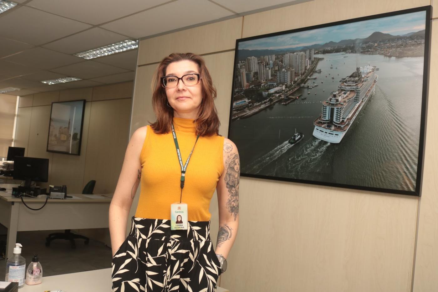 servidora posa ao lado de quadro com navio #paratodosverem