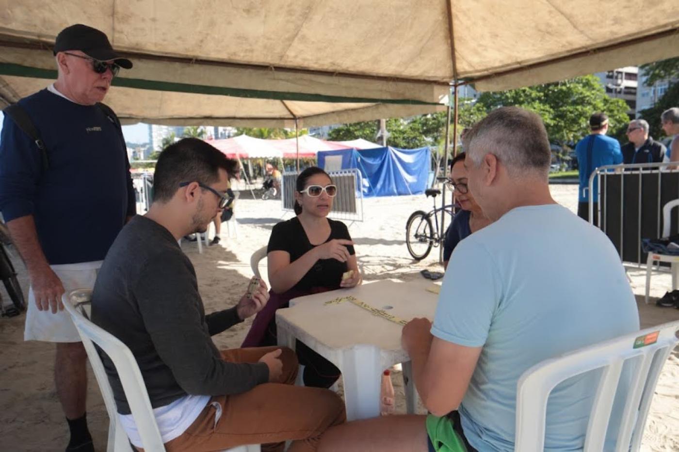 três pessoas estão sentadas à mesa jogando dominó. Um está em pé. Eles estão sob uma barraca na areia da praia. #paratodosverem