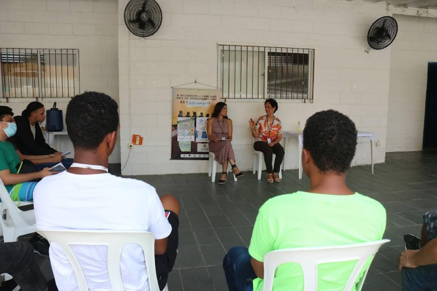 pessoas estão reunidas em um salão. Em primeiro plano, dois rapazes estão de costas para a imagem. Ao fundo, duas mulheres estão sentadas conversando. #paratodosverem