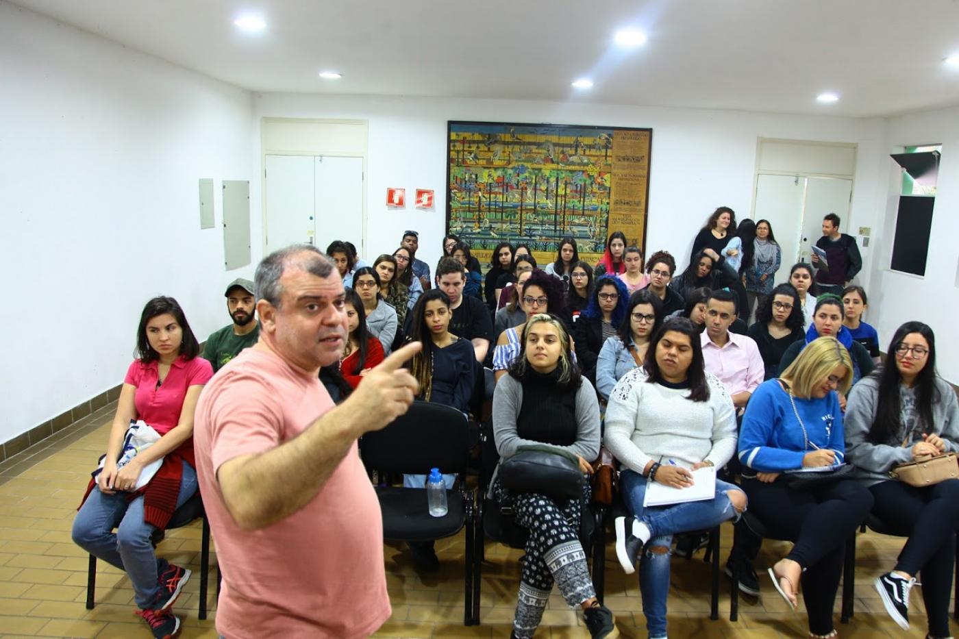 Estudantes estão sentados em auditório e ouvem atentos o palestrante que está à frente de todos. #Pracegover