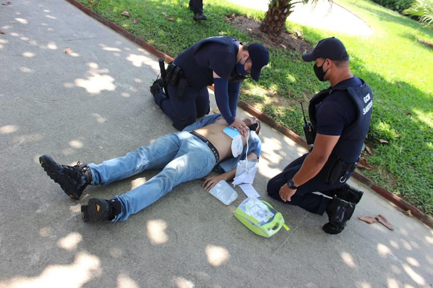 dois guardas estão agachados ao lado de modelo deitado no chão. Um dos guardar coloca o desfibrilador no peito do boneco. #paratodosverem