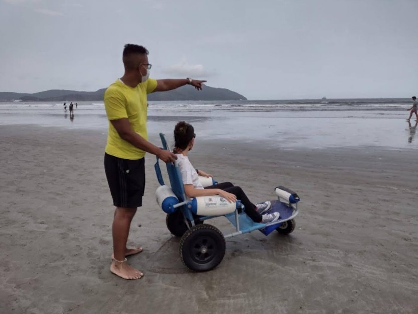 homem segura cadeira própria para entrar no mar onde está sentada uma jovem. ambos estão de costas para a foto.Ele aponta para o mar