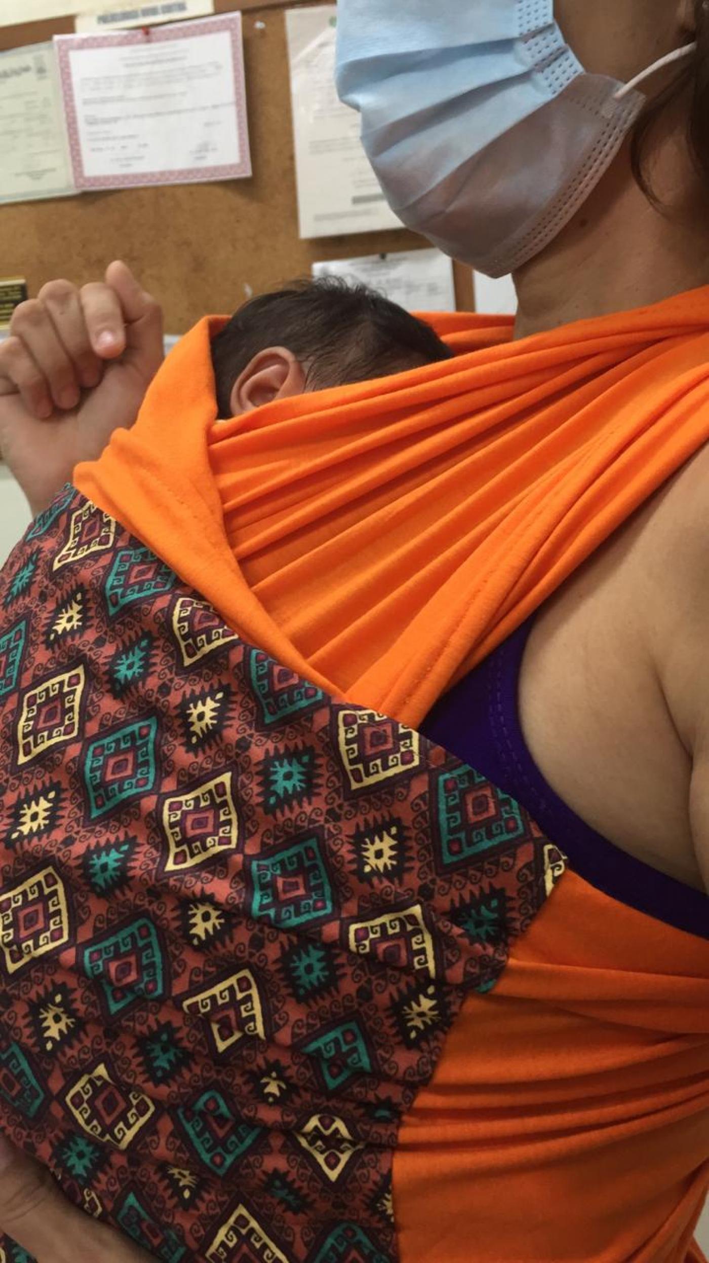 Mulher de máscara usando sling na cor laranja, com recém-nascido junto ao corpo. #pracegover
