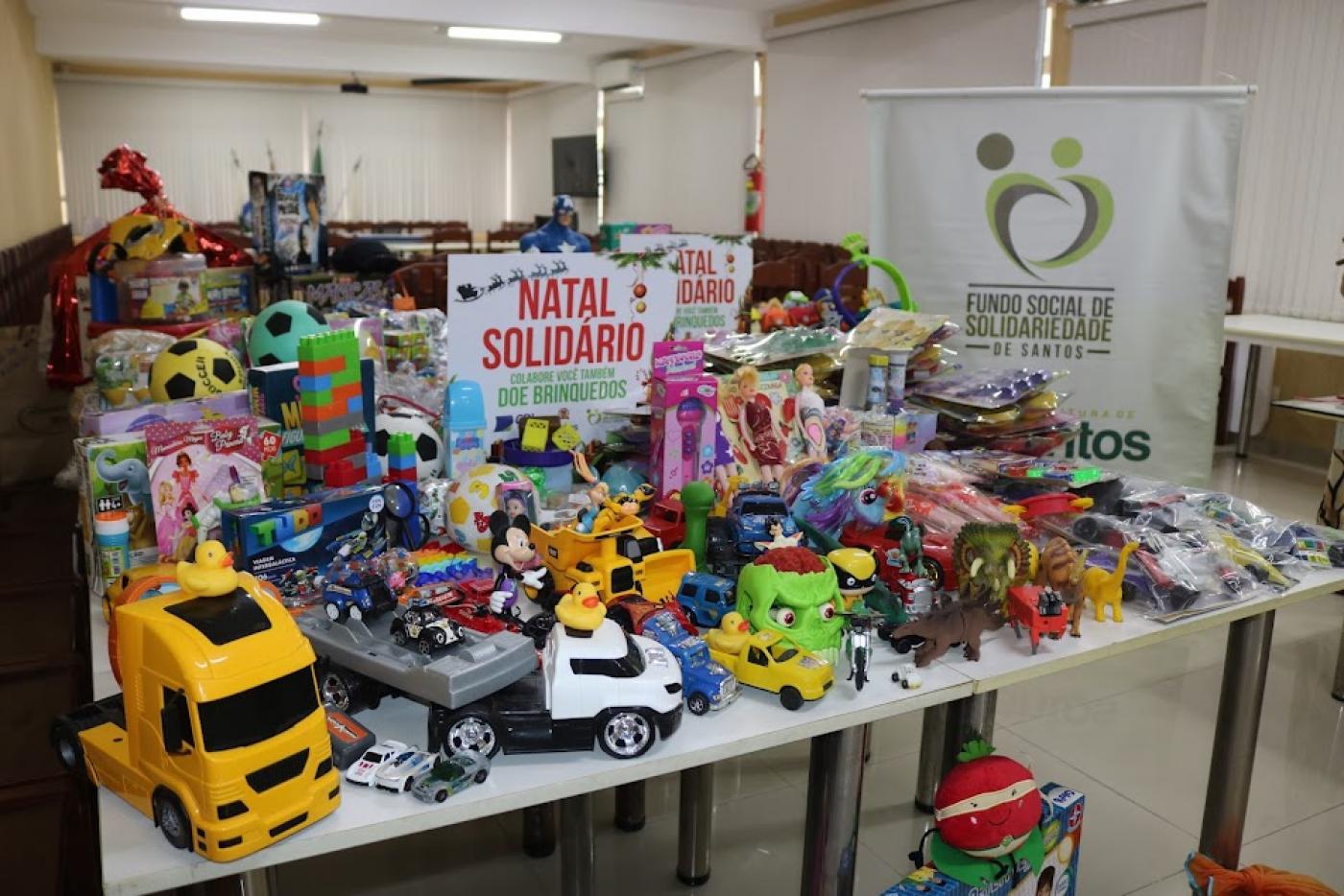 mesa cheia de brinquedos com uma placa no meio onde se lê Natal Solidário. #paratodosverem