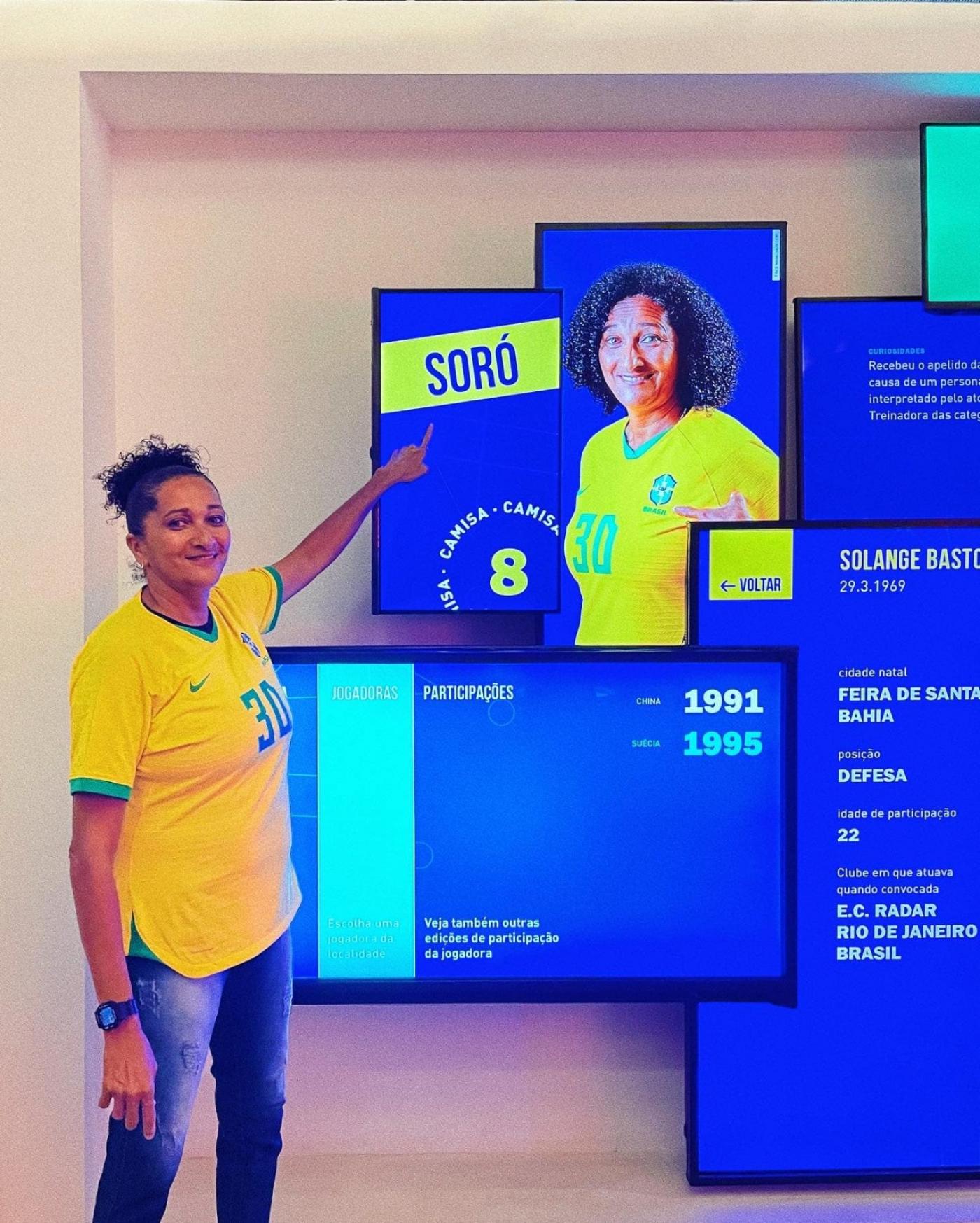 soré está em pé vestindo a camisa da seleção brasileira e apontando para um painel onde aparece a foto dela. #paratodosverem 