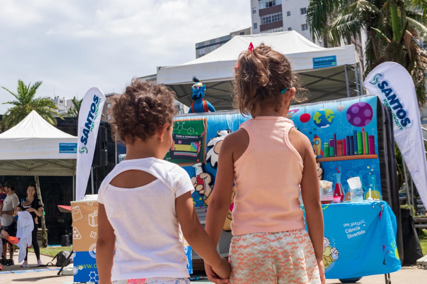 crianças estão de mãos dadas em frente a estande de brincadeiras ao ar livre. #paratodosverem 