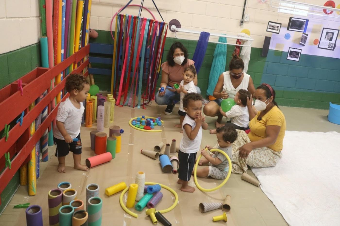 crianças uniformizadas brincam em espaço com  peças coloridas. Elas estão acompanhadas por mulheres que também estão sentadas no chão. #paratodosverem