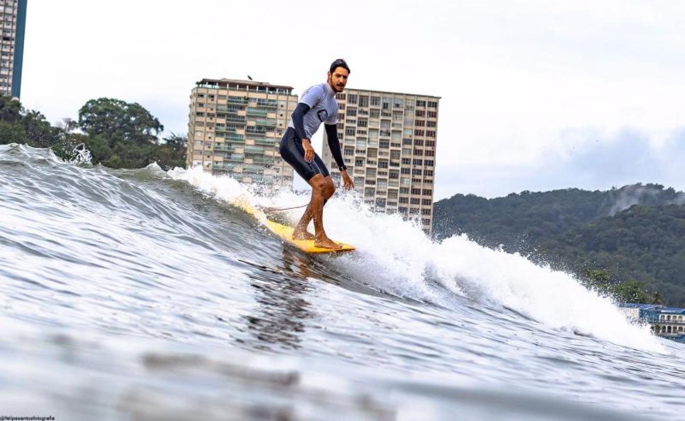 Atleta surfando na crista da onda em prancha longa com prédios e morro ao fundo. #paratodosverem