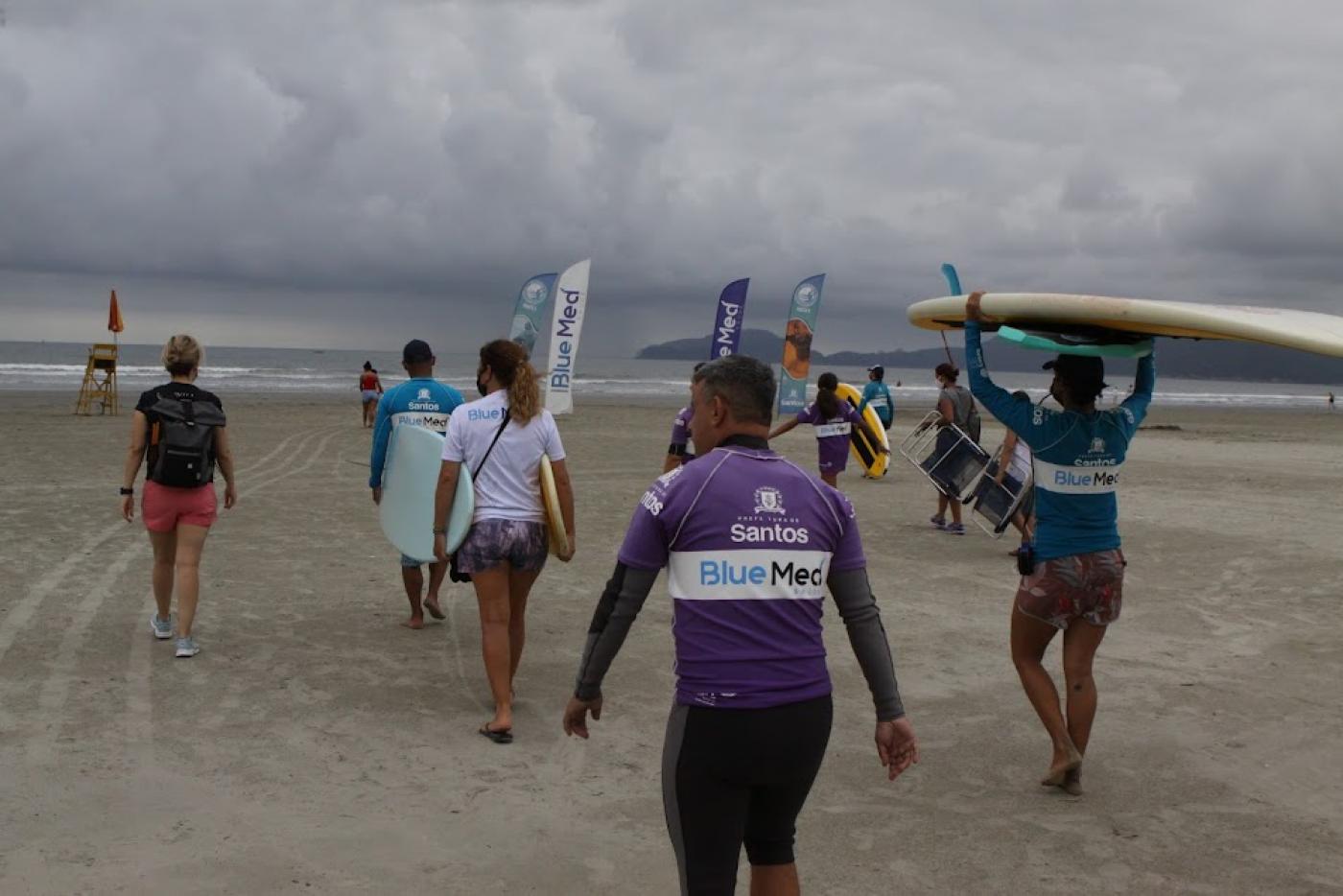 Alunos se dirigem ao mar carregando pranchas no alto da cabeça ou nos braços. O dia está nublado. #paratodosverem