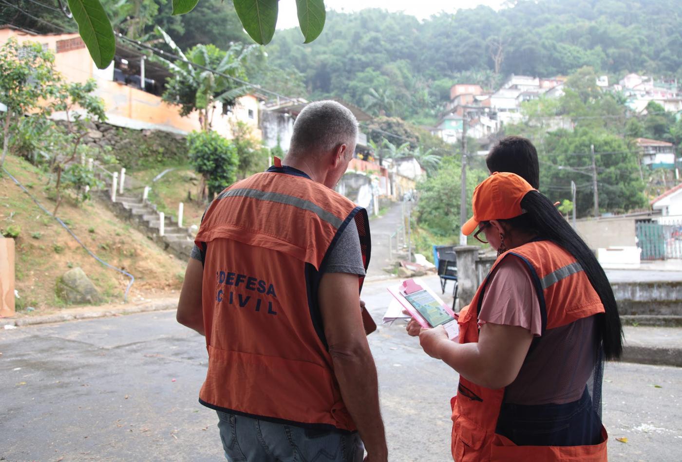 Agentes da Defesa Civil uniformizados e reunidos em rua de morro. #Pracegover