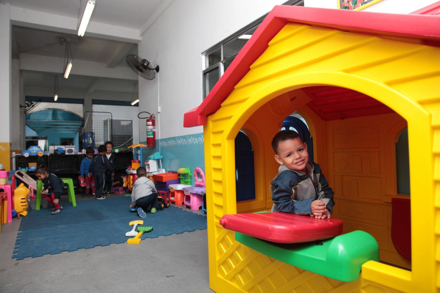 Criança está em casinha de boneca, em primeiro plano. Ao fundo, outras crianças brincam. #Pracegover