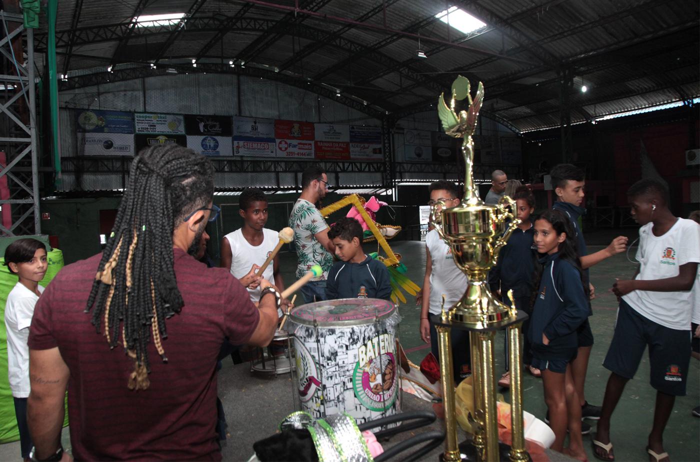 Estudantes estão na quadra da escola de samba em meio a troféus e instrumentos de percussão. #Paratodosverem