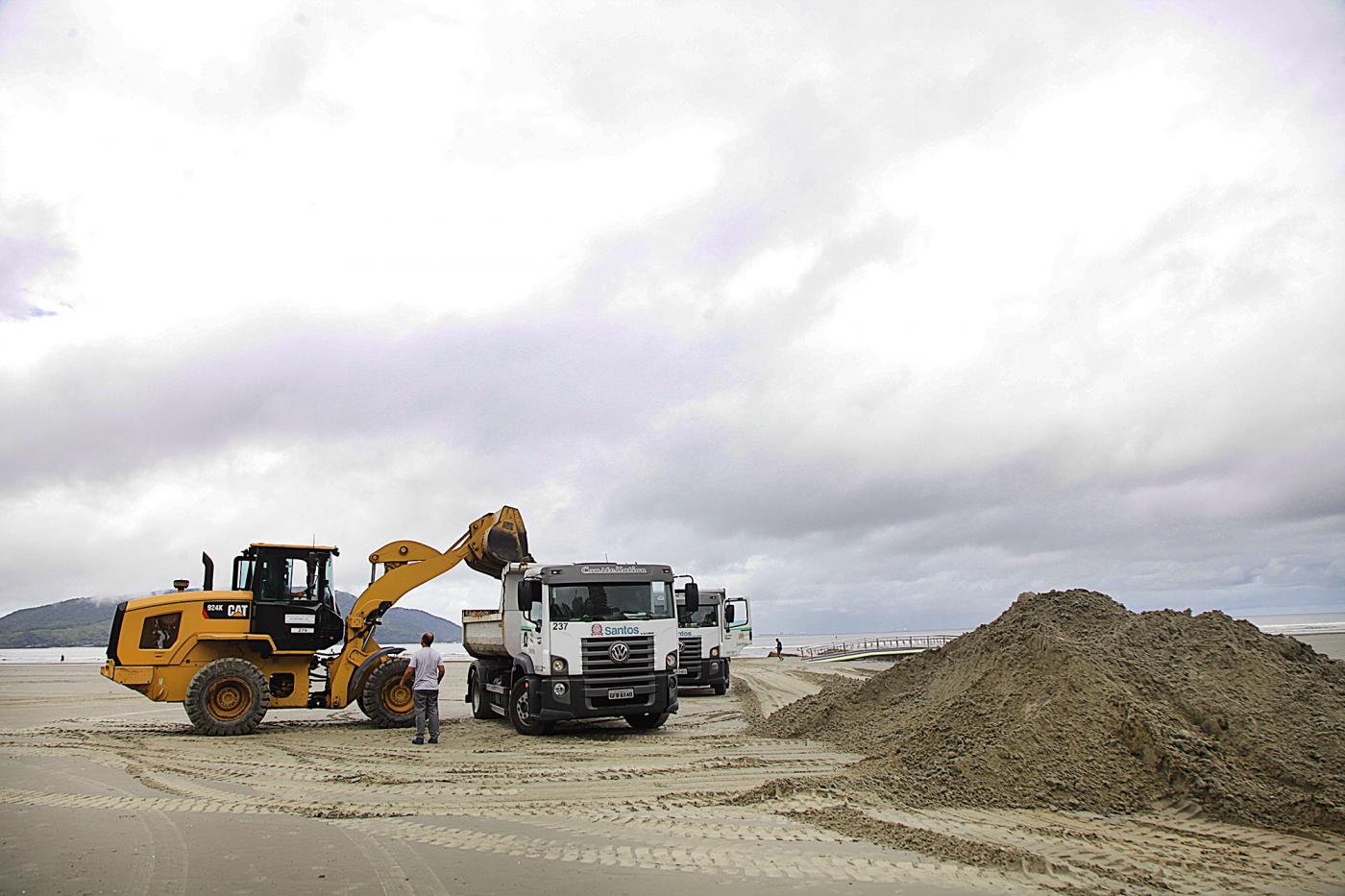 Escavadeira está depositando areia em caminhão. Os dois veículos estão na areia da praia. Ao lado direito da imagem há um monte de areia. #Paratodosverem.