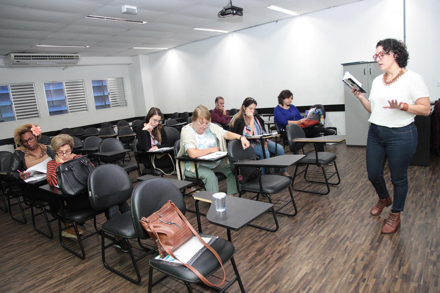 Professora com livro na mão fala para colegas em sala de aula #pracegover 