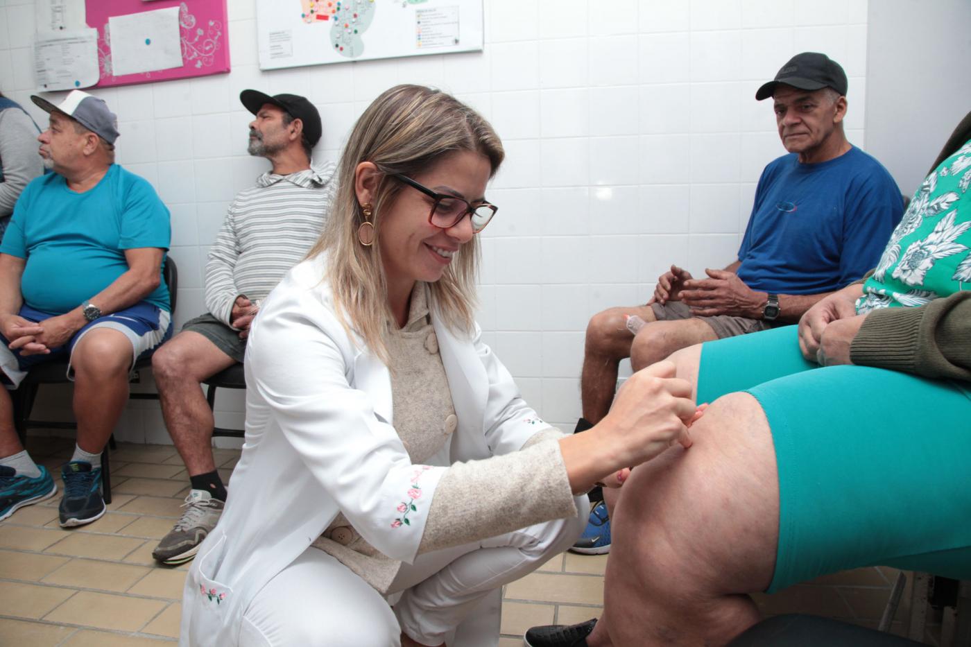 Mulher vestida de roupa branca insere agulhas em joelho de outra mulher, cujo rosto não aparece. Há outras pessoas sentadas no ambiente. #Pracegover