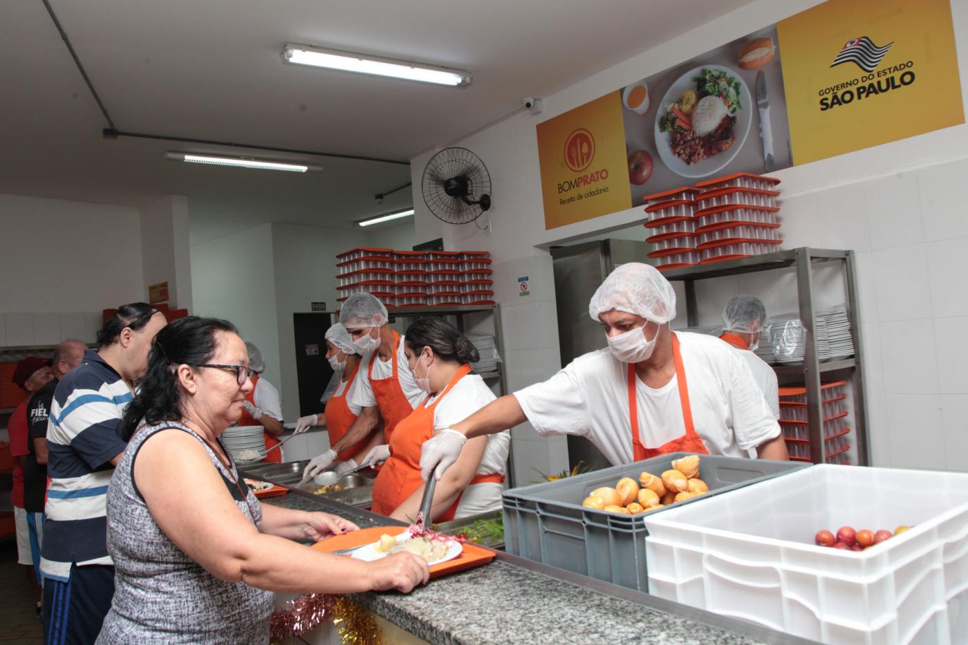 Pessoas estão à frente de balcão segurando bandejas. Do outro lado, pessoas com toucas e máscara servem as que seguram bandejas. #Pracegover