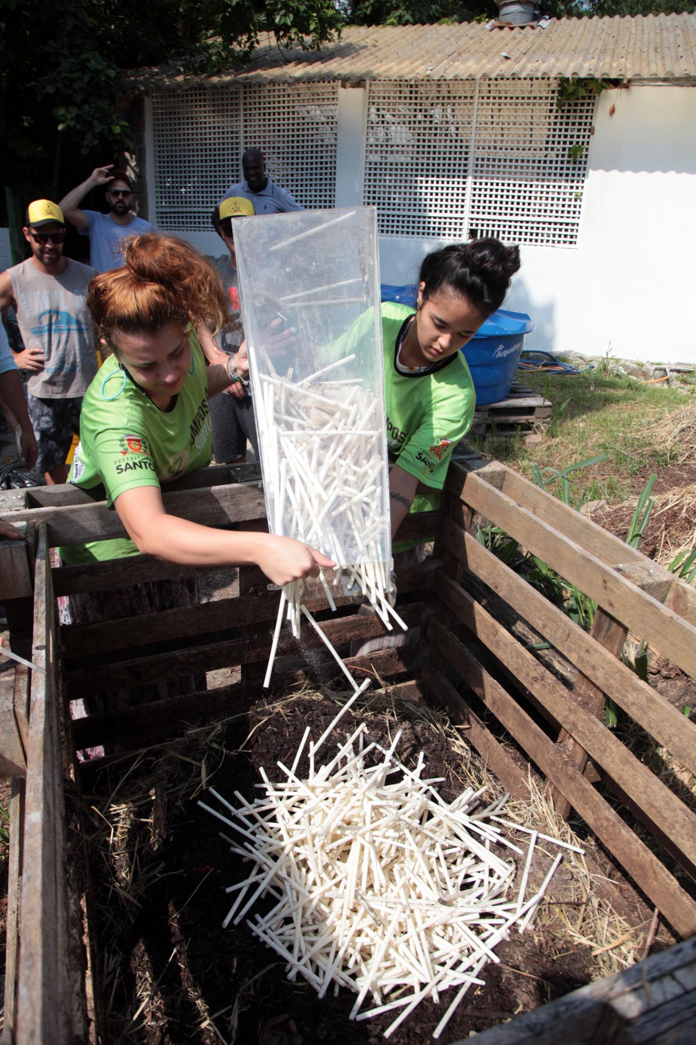 Duas mulheres seguram recipiente e depositam o conteúdo em uma composteira. #Paratodosverem