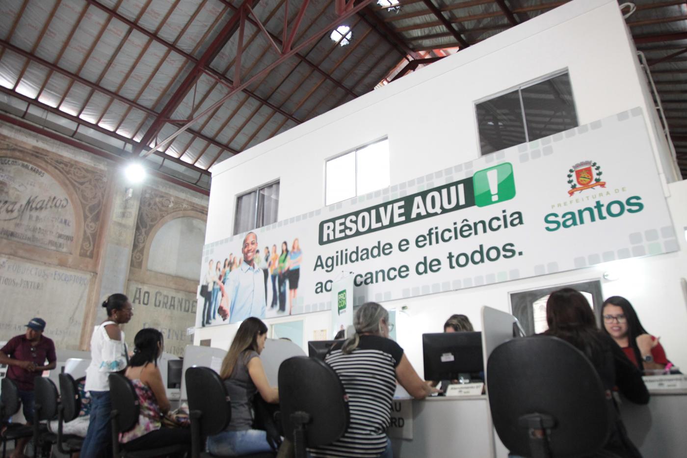 Pessoas estão sentadas diante de balcão e conversam com atendentes. #Pracegover