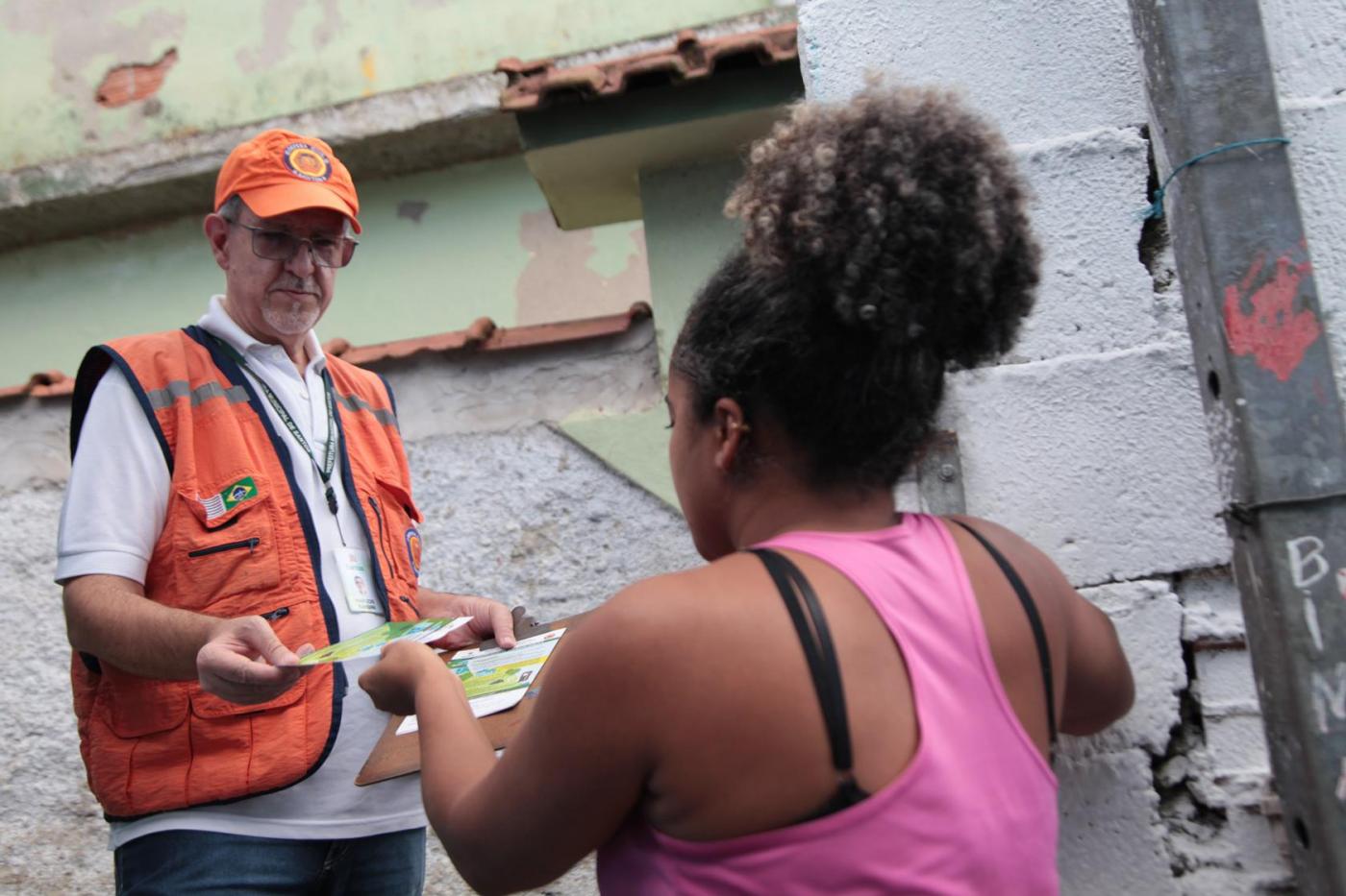 Agente da defesa civil usando colete e boné de identificação entrega folheto para moradores de área de risco. A imagem é fechada. Dá para perceber que os dois estão entre muros. #Paratodosverem