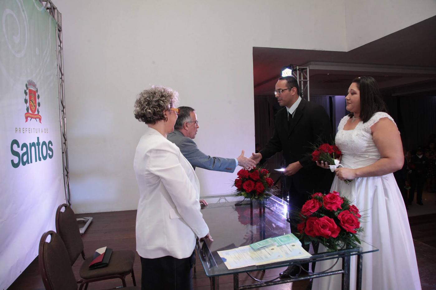 Casal de noivos, com as roupas tradicionais, está na frente de um casal atrás de uma mesa. Ao fundo um painel com o brasão de Santos. #Pracegover