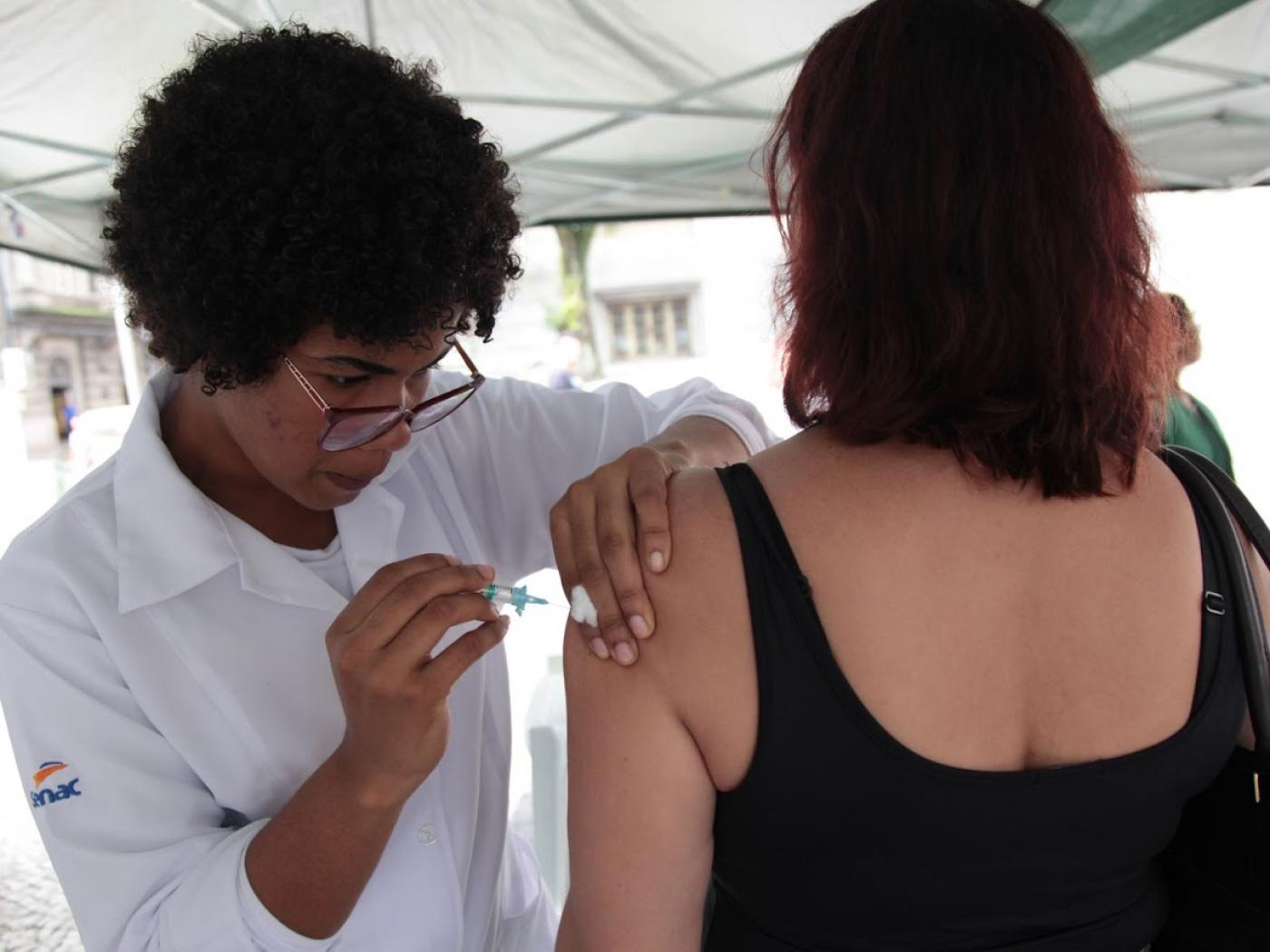 Mulher usando avental branco vacina o braço de outra mulher que está de costas. #Paratodosverem