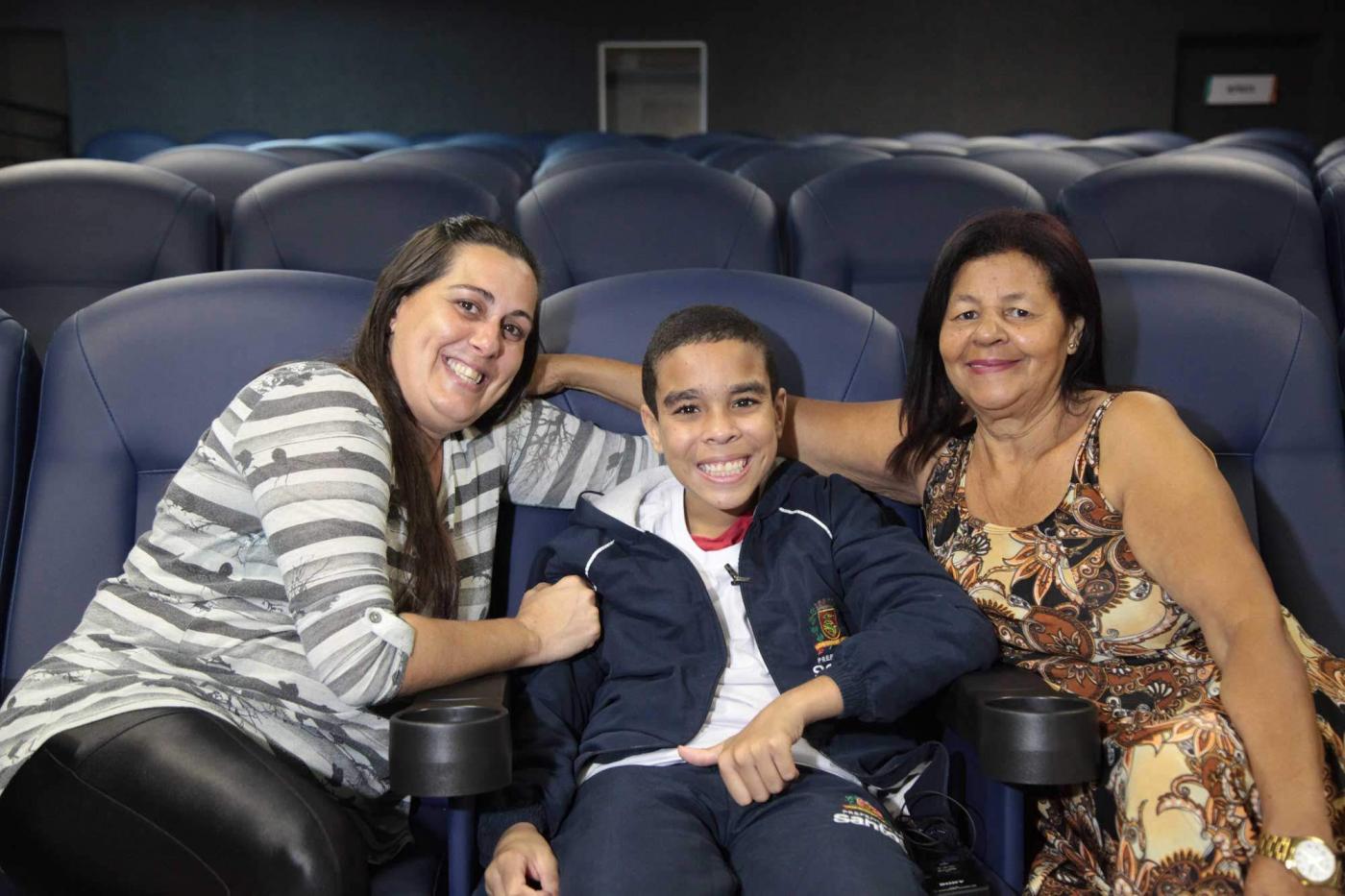 Sentado em cadeira do cinema, Kauê sorri ladeado pela mãe e pela avó
