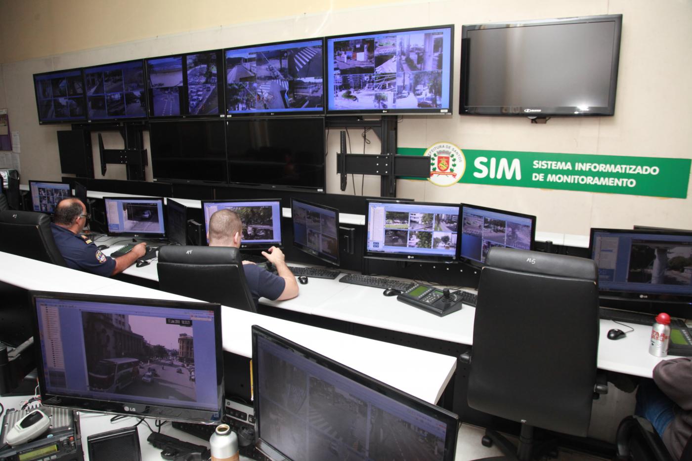 Sala de monitoramento das câmeras distribuídas pela Cidade. Há várias telas distribuídas pela sala. Dois homens estão assistindo. #Pracegover