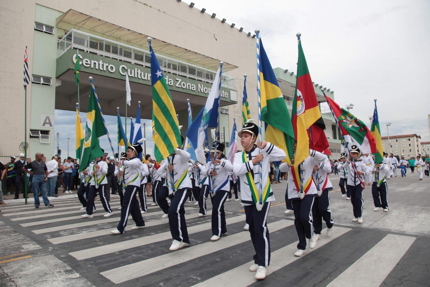 Estudantes uniformizados e segurando bandeiras percorrem avenida. #Pracegover