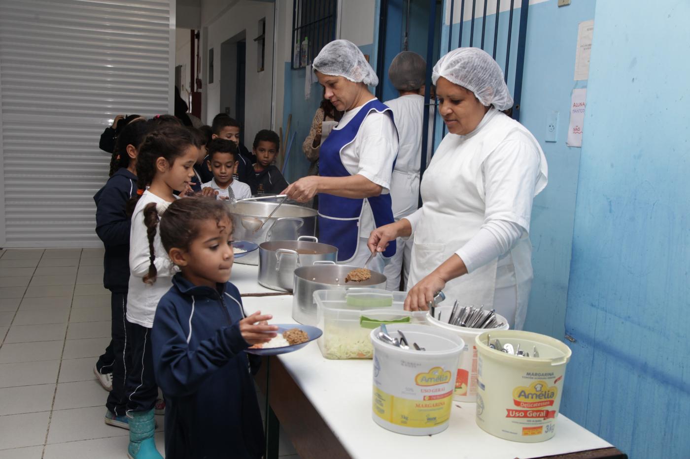 #pracegover Fila de crianças passa por mesa onde duas mulheres com tocas de proteção servem a merenda escolar