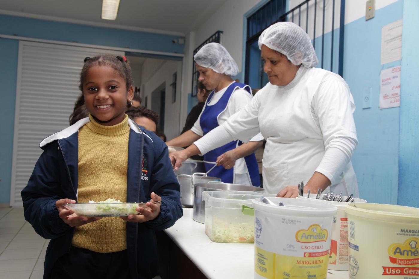 Criança caminha ao lado de mesa onde estão disponíveis os pratos com alimentos. Ela segura um prato com as duas mãos. #Pracegover