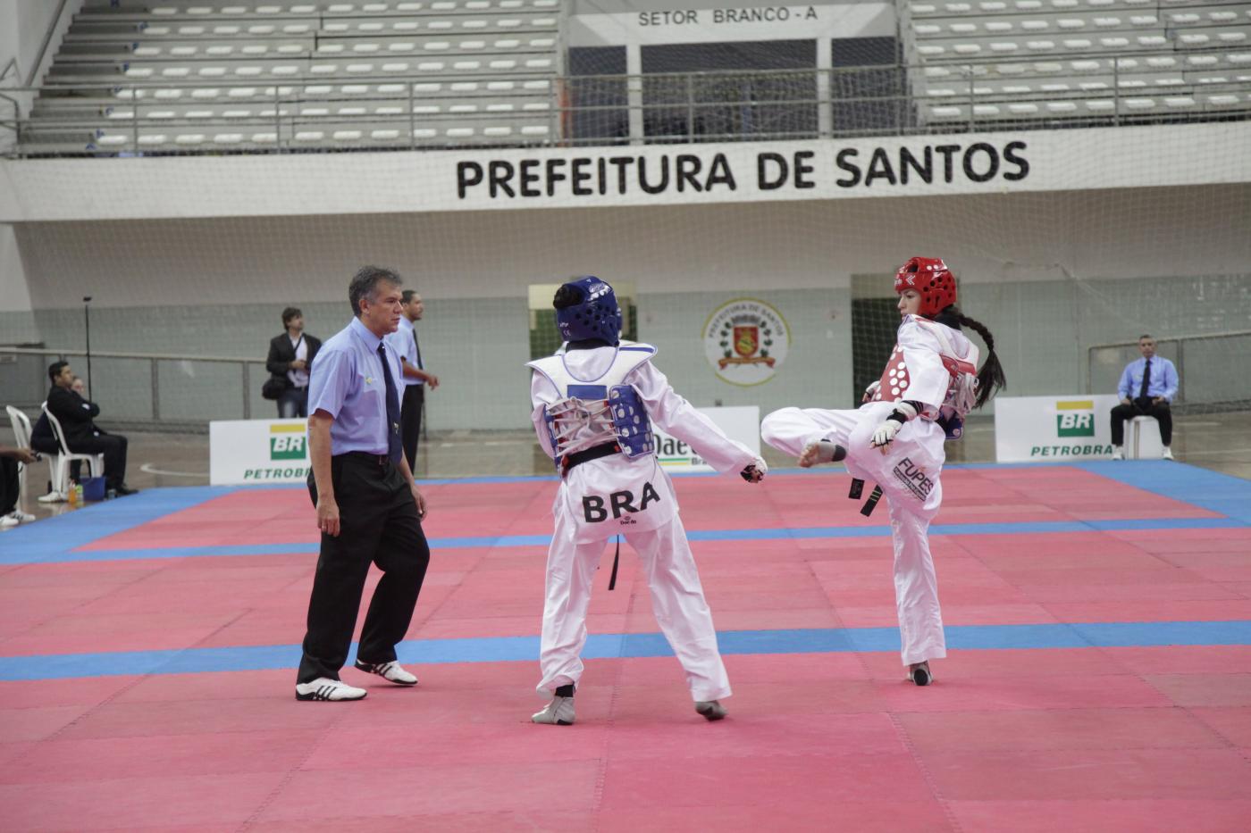 Duas atletas de taekwondo em luta sobre tatame. Ao lado esquerdo está o árbitro. #Pracegover
